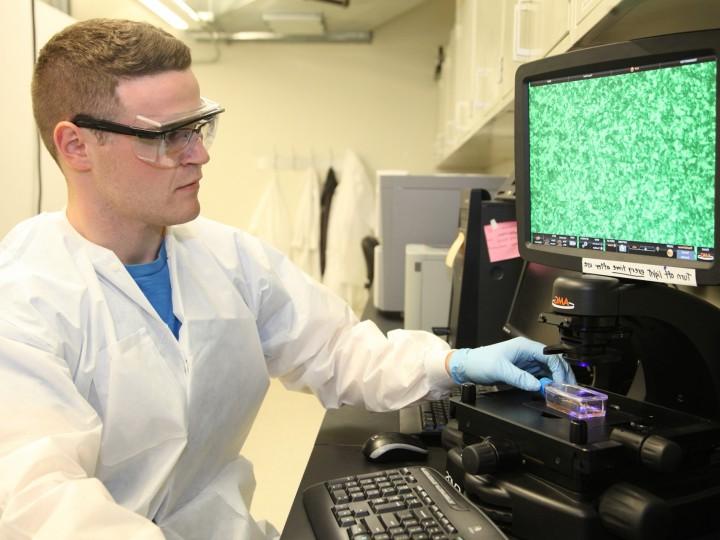 A student wearing safety glasses and white coat studies lab project on laptop